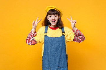 Portrait of cheerful excited girl teenager in french beret, denim sundress showing victory sign isolated on yellow background in studio. People sincere emotions, lifestyle concept. Mock up copy space.