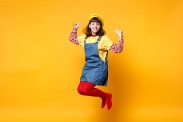 Fototapeta na wymiar Happy girl teenager in french beret, denim sundress showing victory sign jumping and fooling around isolated on yellow wall background in studio. People emotions lifestyle concept. Mock up copy space.