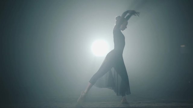 Graceful hard-working ballerina dancing in black dress in the studio in spotlight on a black background. Diligent ballet dancer performing dancing elements of classical ballet. Slow motion.