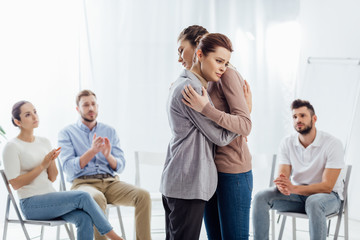 women hugging while group of people sitting and applauding during therapy session