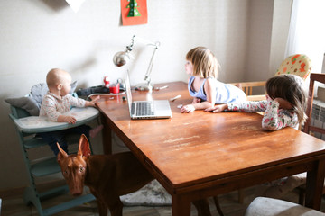 Fototapeta premium baby in highchair and two sisters watch on laptop