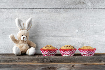 Osterhase aus Stoff, sitzt entspannt winkend mit Muffins vor Holzwand