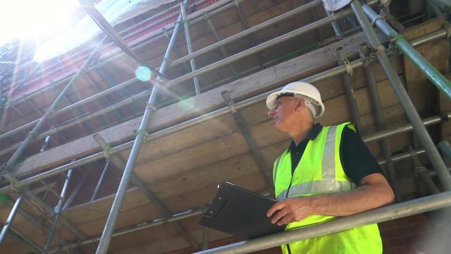 Moving camera clip of male builder foreman, surveyor, construction worker, surveyor, or architect working on construction building site standing on scaffolding writing on clipboard 