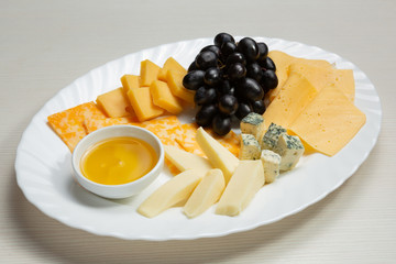 five sorts of cheese are laid out on a plate, cut into pieces, a bunch of grapes and honey in a bowl lie next to it, an assortment of cheese
