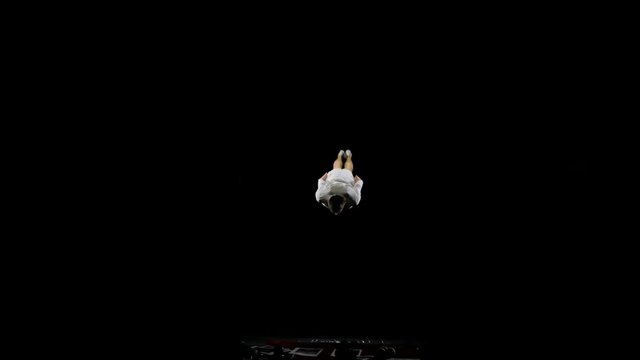 Athletic man in white clothes performs tricks jumping on a trampoline on a black monophonic background