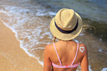 Pretty young tanned girl in straw hat and swimsuit with sunscreen on her shoulder relaxing near sea in family vacation in sunny summer day. Healthcare. Skincare. Leisure. Childhood