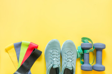 Sport equipment, rubber band, dumbbells, fitness shoes, measuring tape on yellow.
