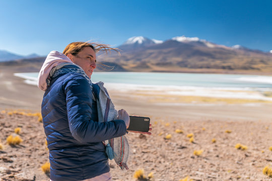 Landscape woman photographer taking mobile phone photos in an amazing wilderness environment at Atacama Desert Andes mountains lagoons. A woman cut out silhouette over the awe Tuyajto Lagoon scenery
