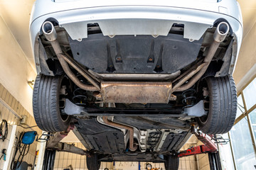 Car chassis on the lift, view from the bottom. Visible exhaust system, wheels, brake hoses. - obrazy, fototapety, plakaty