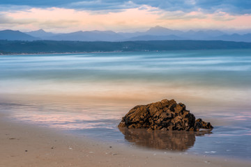 Dreamy waves breaking in slow motion at Plettenberg Bay beach at sunset, with hills in the distance and a rock in the foreground reflected in the water. Garden Route, Western Cape, South Africa