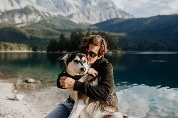 Mensch und Hund umarmen sich an einem schönen See mit Bergen im Hintergrund - Freundschaft