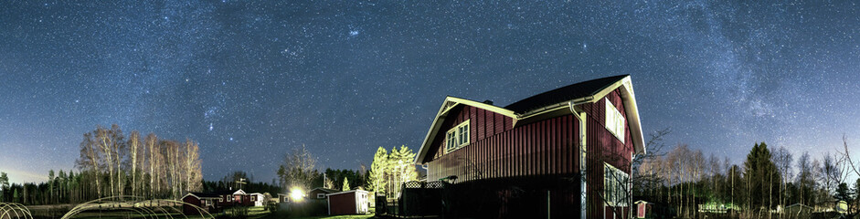 Panorama of very clear deep starry skies with Milky Way above traditional Scandinavian wooden red house at countryside