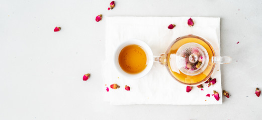 Top view of two white cups and transparent teapot with herbal tea of dried pink roses buds over textile napkin on gray background with copy space. Long wide banner.