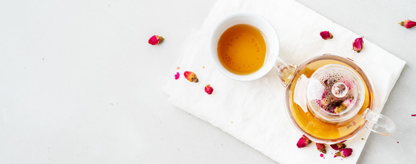 Top view of two white cups and transparent teapot with herbal tea of dried pink roses buds over textile napkin on gray background with copy space. Long wide banner.