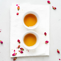 Top view of two white cups and transparent teapot with herbal tea of dried pink roses buds over textile napkin on gray background with copy space. Brewing and Drinking tea.