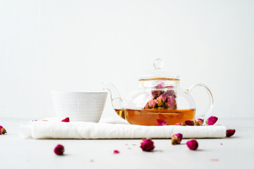 White teacup and transparent teapot with herbal tea of dried pink roses buds on white background with copy space. Brewing and Drinking tea.