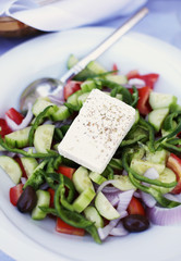 Delicious traditional Greek salad with tomatoes, cucumbers, pepper, olives, green salad, spices and feta cheese served on a white plate.