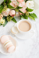 Elegant sweet dessert macarons, cup of coffee and pastel colored beige flowers bouquet on white marble
