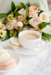 Elegant sweet dessert macarons, cup of coffee and pastel colored beige flowers bouquet on white marble