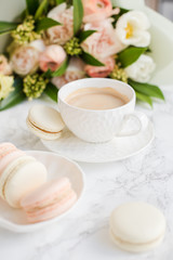 Elegant sweet dessert macarons, cup of coffee and pastel colored beige flowers bouquet on white marble