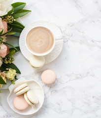 Elegant sweet dessert macarons, cup of coffee and pastel colored beige flowers bouquet on white marble