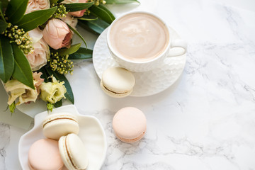 Elegant sweet dessert macarons, cup of coffee and pastel colored beige flowers bouquet on white marble