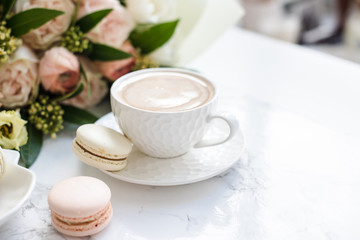 Elegant sweet dessert macarons, cup of coffee and pastel colored beige flowers bouquet on white marble