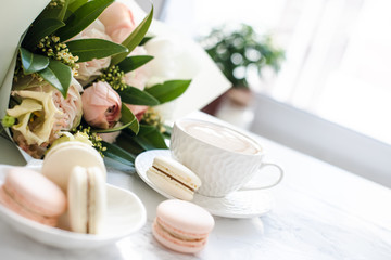 Elegant sweet dessert macarons, cup of coffee and pastel colored beige flowers bouquet on white marble