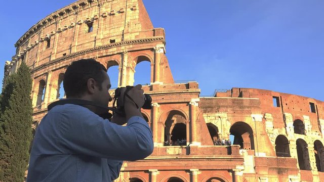 Man with back take pictures to Colosseum, blue serene sky in background, iconic ancient Roman gladiatorial arena,  the largest amphitheatre ever built