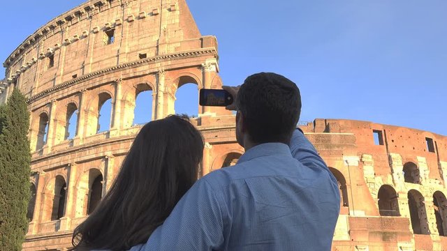 Couple with back take pictures with phone to Colosseum in Rome, blue serene sky