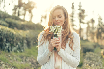 Happy woman with red long hair and a pleasant smile in the rays of the sun holds a bouquet of delicate flowers in front of her and enjoys