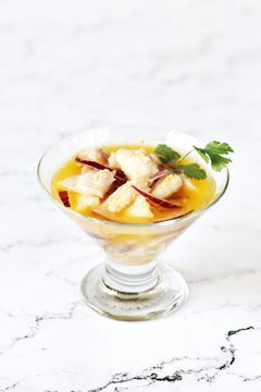 white fish ceviche in orange juice, served in a glass ice-cream bowl against a background of white marble. raw fish is marinated in lemon juice and served with onions, citrus juice and greens. 
