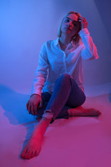 Beautiful girl fashion model sitting down in studio with colorful blue and red light. Woman wears white shirt, blue jeans and with bare feet. Low key light setup