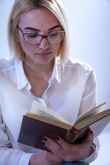 Student girl reading old book with glasses. Young blonde women wears white elegant shirt. White background in studio. Close up, selective focus