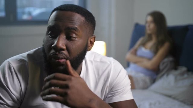 Close up african american male failed in bed grabbing head due to potency problems at rather young age. Blurry on background needing love disappointed female suffering from impotence of man