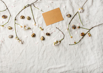 Easter holiday Spring mood flat-lay with equal eggs and blooming branches of almond tree with white flowers over light gray linen tablecloth, top view, copy space on blank craft paper card