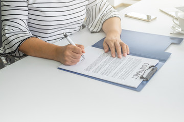A women hands signing contract in blue folder on top of white table with bright light