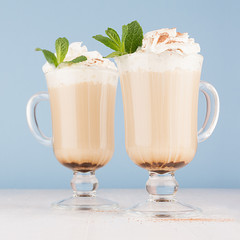 Coffee beverage with whipped cream, fresh green mint, cocoa powder in pastel blue interior on white wooden table, square, closeup.