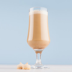 Macchiato coffee with layers, cream and cookies in transparent goblet on pastel blue background and white wooden table, square.