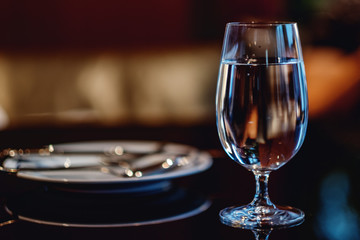 Glass of fresh mineral water on table in cafe or restaurant. Empty ceramic plate on dark background in blur. Horizontal image with selective focus on the glass and copy space for text. 