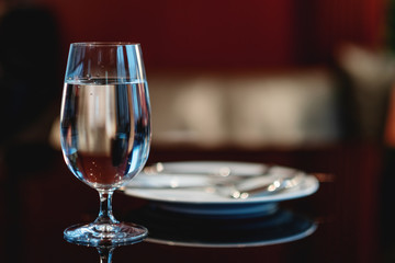 Glass of fresh mineral water on table in cafe or restaurant. Empty ceramic plate on dark background in blur. Horizontal image with selective focus on the glass and copy space for text. 