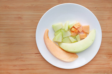 Fresh sliced of green and orange cantaloupe melon on white plate against wooden board background with copy space.