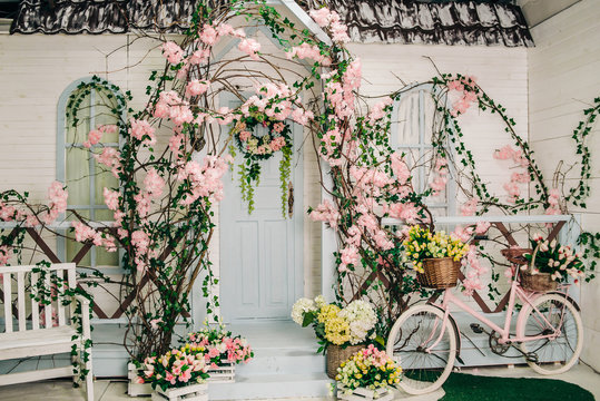 the facade of a white building with a porch decorated with blooming flowers, baskets of flowers next to the steps and a pink bike, decorations for a photo studio in spring