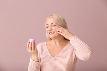 Mature woman applying facial cream on color background