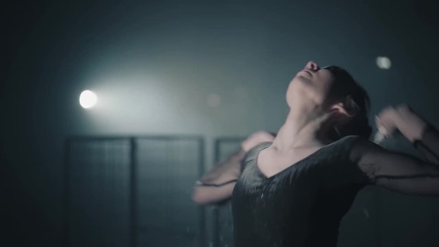 Portrait of professional ballerina dancing in black dress in the studio in spotlight on black background. Young beautiful ballet dancer arching body back and throwing flour. Slow motion.