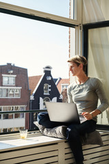 Woman with short hair sitting at window with canal view sill with laptop and coffee, working on a project in private apartment.