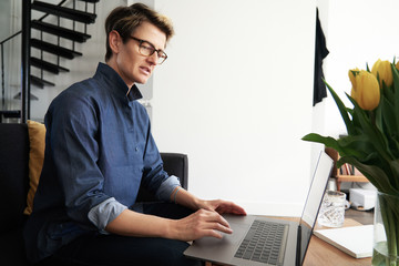 Business woman with short hair and glasses working on laptop at modern apartment, opened window, sunny daylight. Concept of young entrepreneur works on start-up..