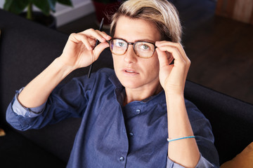 Portrait of business woman with short hair looks to camera into glasses sitting at modern apartment. Daylight.