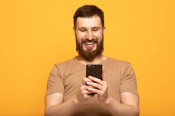 Close up shot portrait of young bearded man isolated on yellow background, looking agitated at display of her smartphone smiling and laughing happily, impressed by media content from web