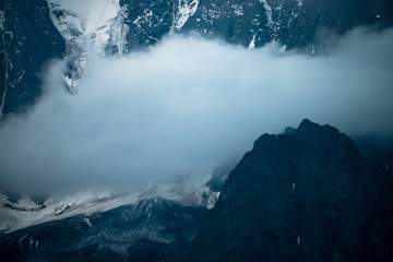 Low cloud before huge glacier. Giant snowy rocky mountain wall in thick fog. Early morning in mountains. Impenetrable fog. Dark atmospheric foggy landscape with cold rocks. Tranquil mystic atmosphere.